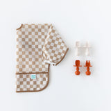 Bumkins Sleeved Bib: Sand Check lays folded next to two sets of silicone utensils on a white background.