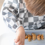 Close-up of a child practicing baby-led weaning, enjoying small pancakes while wearing a Bumkins Sleeved Bib: Charcoal Check.