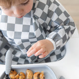 A child in a Bumkins Sleeved Bib: Charcoal Check reaches for pancakes, embracing baby-led weaning at a white table.