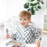 A child in a Bumkins Sleeved Bib: Charcoal Check sits in a high chair, smiling with a spoon, ready for baby-led weaning fun.
