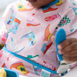 A child in a vibrant Bumkins Sleeved Bib: Boots grips a blue utensil by a blue bowl, ideal for baby-led weaning.