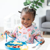 A toddler in a Bumkins Sleeved Bib: Boots sits in a high chair, smiling while eating from a blue plate with matching utensils.