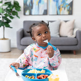 A toddler enjoys baby-led weaning from a blue plate, wearing Bumkins Sleeved Bib: Boots in front of a gray sofa, potted plant, and art.