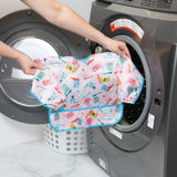 A person holds Bumkins Sleeved Bib: Boots in front of an open front-loading washer, with a laundry basket nearby.