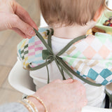 Adult hands tie the ribbon closure of a Bumkins Sleeved Bib: Motley Check around a child's neck.