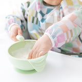 Close-up of a child wearing a Bumkins Sleeved Bib: Motley Check reaching into a green bowl while holding a spoon.