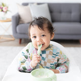 A toddler wears a Bumkins Sleeved Bib: Motley Check while sitting in a high chair, chewing a green spoon near a green bowl.