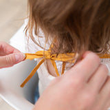 Adult hands adjust the ribbon closure of a bib from Bumkins On-The-Go Mealtime 6-Piece Set: Wander + Sand around a child's neck.