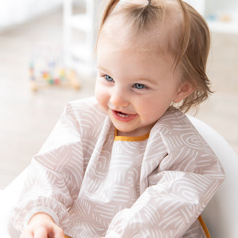 A blue-eyed girl smiles in a high chair, wearing a bib from Bumkins On-The-Go Mealtime 6-Piece Set: Wander + Sand.