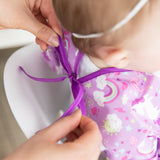 An adult ties the ribbon closure of a Bumkins Sleeved Bib: Unicorns around a child's neck.