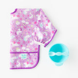 Bumkins Sleeved Bib: Unicorns lays folded next to a blue bowl & spoon set on a white background.
