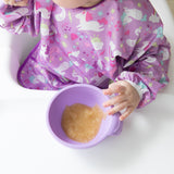 A baby in a high chair wearing a Bumkins Sleeved Bib: Unicorns eagerly holds a lavender bowl of applesauce.