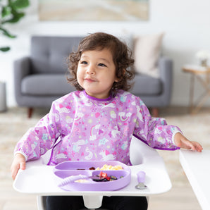 A child in a Bumkins Sleeved Bib: Unicorns smiles in a high chair, enjoying food from a purple divided plate with matching utensils.