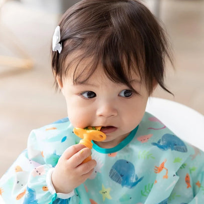 A toddler wearing a colorful bib with ocean animals is eating with a yellow spoon. The child has a hair clip on one side and looks thoughtfully to the side while holding the spoon to their mouth.