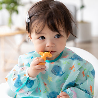 A girl in a Bumkins SuperBib® and Sleeved Bib 3-Piece Set: Rolling With The Waves, Whale Tail, Ocean Life chews an orange spoon.