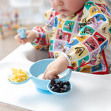 A child wearing Bumkins Sleeved Bib: Mickey & Friends Check Mates eats from a blue bowl with cheese and blueberries on a highchair.