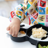 A child in Bumkins Sleeved Bib: Mickey & Friends Check Mates enjoys baby-led weaning from a divided plate with macaroni at the table.