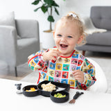 A toddler with a ponytail smiles while eating from a black plate on a white table, wearing Bumkins Sleeved Bib: Mickey & Friends Check Mates.