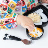 A child in Bumkins Sleeved Bib: Mickey & Friends Check Mates enjoys baby-led weaning from a Mickey-shaped plate.