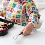 A child in Bumkins Sleeved Bib: Mickey & Friends Check Mates holds a spoon at a white table in front of a plate with raspberries.