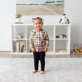 A toddler in a Bumkins Sleeved Bib: Mickey & Friends Check Mates smiles before a toy-filled shelf in a cozy room.