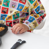 A child in Bumkins Sleeved Bib: Mickey & Friends Check Mates sits at a white table with a black dish and utensil.