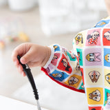Baby's hand holding a spoon, wearing Bumkins Sleeved Bib: Mickey & Friends Check Mates in a bright room.