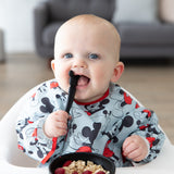 A baby in a high chair wearing a blue bib eats oatmeal and berries from a Bumkins Silicone First Feeding Set: Mickey Mouse Classic Black.