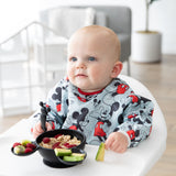 A baby in a high chair wearing a bib eats oatmeal, berries, & avocado from a Bumkins Silicone First Feeding Set: Mickey Mouse Classic Black.