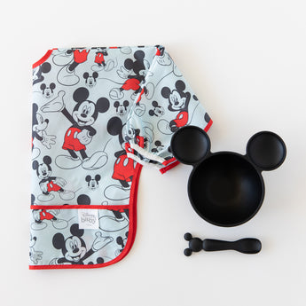 Bumkins Sleeved Bib: Mickey Mouse lays folded next to a black Mickey Mouse-shaped bowl and matching spoon on a white background.