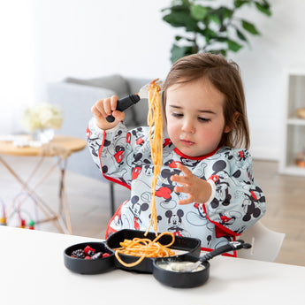 A toddler wearing a Bumkins Sleeved Bib: Mickey Mouse eats spaghetti with a fork from a Mickey Mouse-shaped plate at a white table.