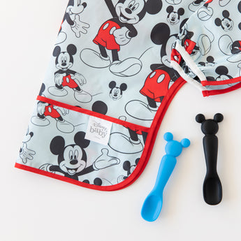 Close-up of Bumkins Sleeved Bib: Mickey Mouse folded on a white background next to 2 spoons with Mickey-shaped handles on a white background.