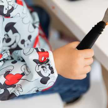 Close-up of a toddler in a Bumkins Sleeved Bib: Mickey Mouse holding a black-handled utensil.