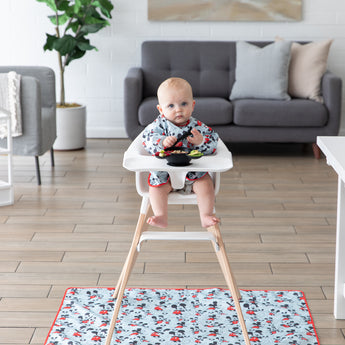 A baby in a Bumkins Sleeved Bib: Mickey Mouse eats from a black bowl in a high chair with a matching mat on the floor underneath.