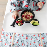 A baby in a high chair wearing a blue bib eats oatmeal & fruit from Bumkins Silicone First Feeding Set: Mickey Mouse Classic Black.
