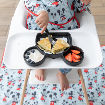 A child in a Bumkins Sleeved Bib: Mickey Mouse sits in a high chair with a matching mat on the floor, eating from a Mickey-shaped plate.