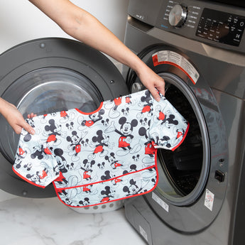 A person holds Bumkins Sleeved Bib: Mickey Mouse in front of an open front-loading washing machine.
