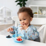 A young boy enjoys self-feeding with Bumkins On-The-Go Mealtime 5-Piece Set: Mickey + Minnie Mouse Sunny Side Up Blue at a white table.