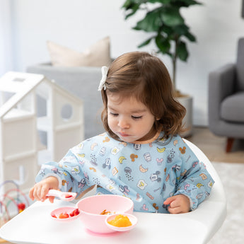 A young girl in a high chair enjoys self-feeding with Bumkins On-The-Go Mealtime 5-Piece Set: Mickey + Minnie Mouse Sunny Side Up Pink.