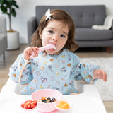 A child in a Bumkins Sleeved Bib: Mickey + Minnie Sunny Side Up enjoys fruit with a spoon, embracing baby-led weaning in a high chair.