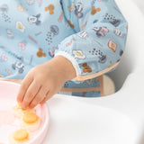 A child's hand reaches for food on a pink tray, wearing Bumkins Sleeved Bib: Mickey + Minnie Sunny Side Up.