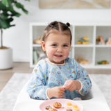 A child with pigtails smiles in a Bumkins Sleeved Bib: Mickey + Minnie Sunny Side Up in a high chair with snacks on a pink plate.