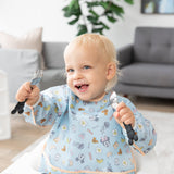 A blonde toddler with a fork and spoon sits in a bright room, wearing Bumkins Sleeved Bib: Mickey + Minnie Sunny Side Up.