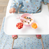 A child in a Bumkins Sleeved Bib: Mickey + Minnie Sunny Side Up enjoys yogurt, berries, granola with strawberries, and orange slices.