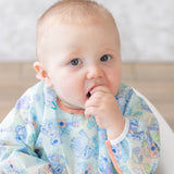 A baby in a Bumkins Sleeved Bib: Aloha Stitch sits curiously, hand in mouth, the waterproof fabric gleaming with colorful patterns.