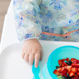 A child's hand wearing a Bumkins Sleeved Bib: Aloha Stitch with a blue spoon and a blue bowl of berries on a high chair tray.