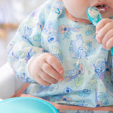 A baby enjoys self-feeding with a spoon, wearing a Bumkins Sleeved Bib: Aloha Stitch made of waterproof fabric.