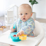 A baby in a high chair, focused on baby-led weaning, wears a charming Bumkins Sleeved Bib: Aloha Stitch against a bright backdrop.