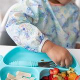 A child in a Bumkins Sleeved Bib: Aloha Stitch practices baby-led weaning with fruit and chicken from a blue divided plate using a fork.