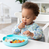 A toddler in a Bumkins Sleeved Bib: Aloha Stitch explores baby-led weaning, enjoying fruit and cheese from a divided blue plate on a white table.